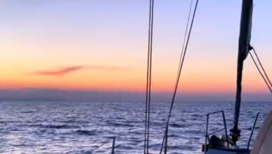 Vista dal ponte di una barca a vela al tramonto, con cielo arancione e rosa e mare increspato.