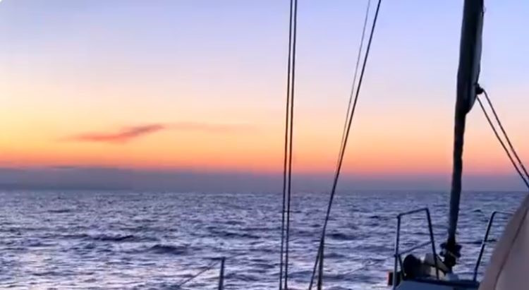 Vista dal ponte di una barca a vela al tramonto, con cielo arancione e rosa e mare increspato.
