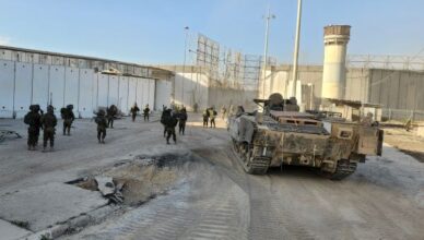 "Un veicolo militare corazzato cingolato si trova su una strada sterrata accanto a alte mura di cemento, con diversi soldati in uniforme ed equipaggiamento completo